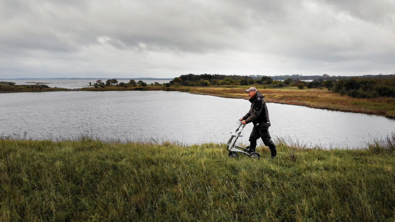 Gebruiker wandelt met byAcre Carbon Overland lichtgewicht rollator langs water in natuurlijke buitenomgeving